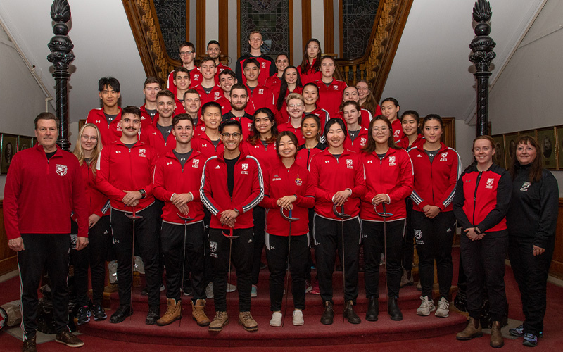 Both men's and women's fencing teams along with the coaching staff on the stairwell in MacKenzie with the front row holding swords.