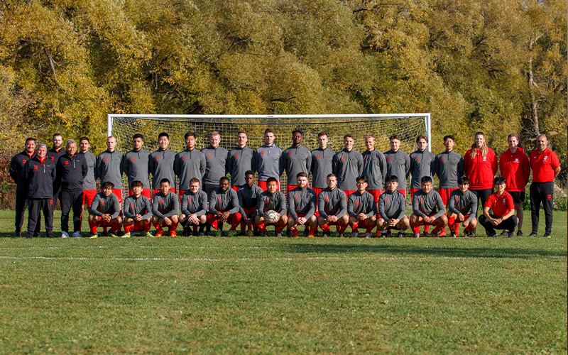 The men's soccer team and support staff/coaches standing in two rows in front of the goal.