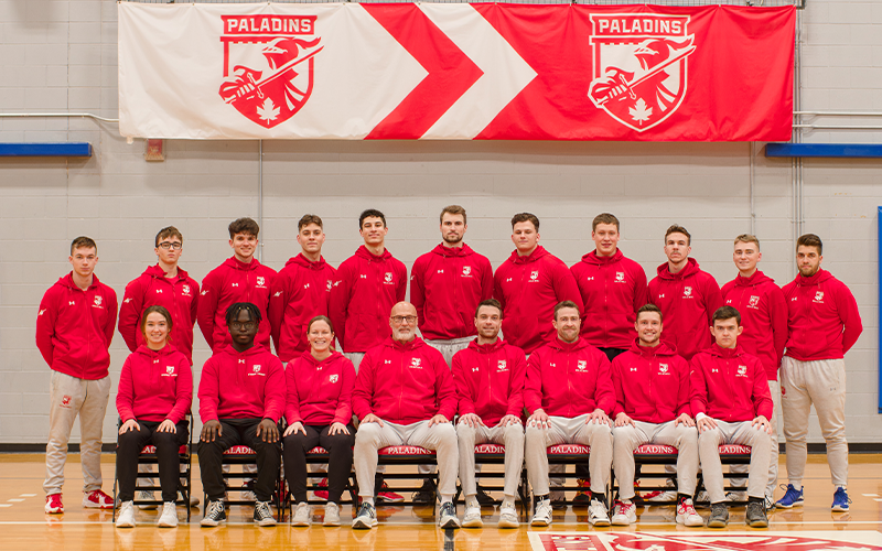14 joueurs de volleyball masculin, leur personnel d'encadrement et leurs entraîneurs devant la bannière des Paladins.