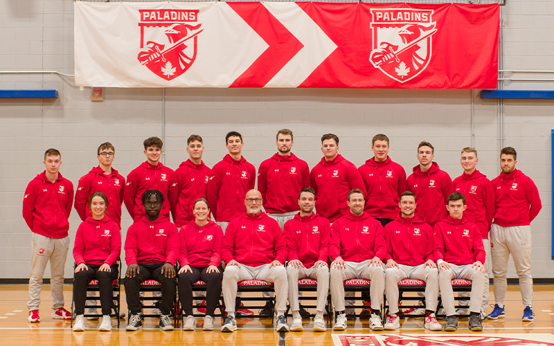 14 men's volleyball players along with their support staff and coaches in front of the Paladins banner.